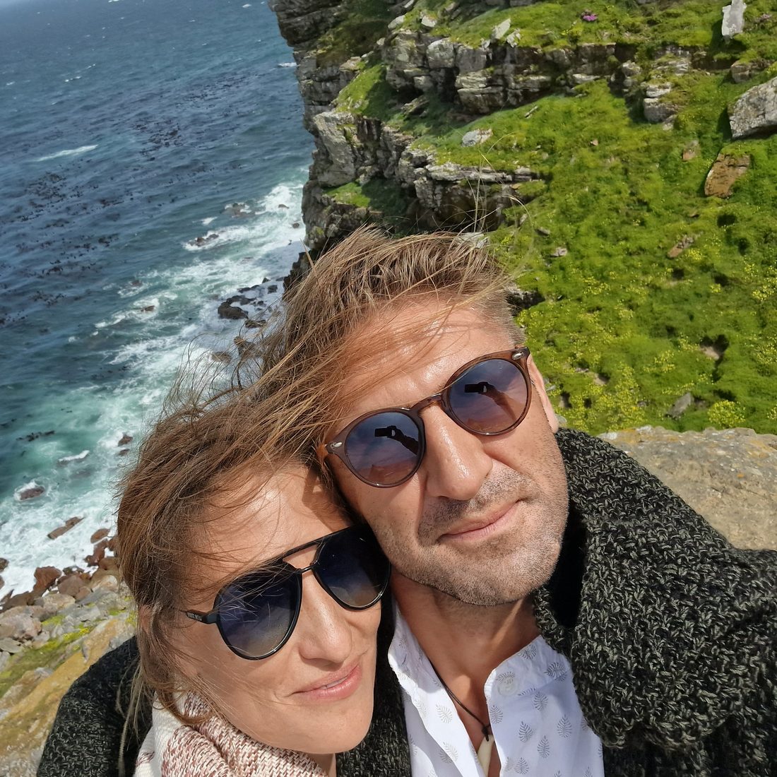 Two of the hosts in sunglasses on a windy clifftop above the sea