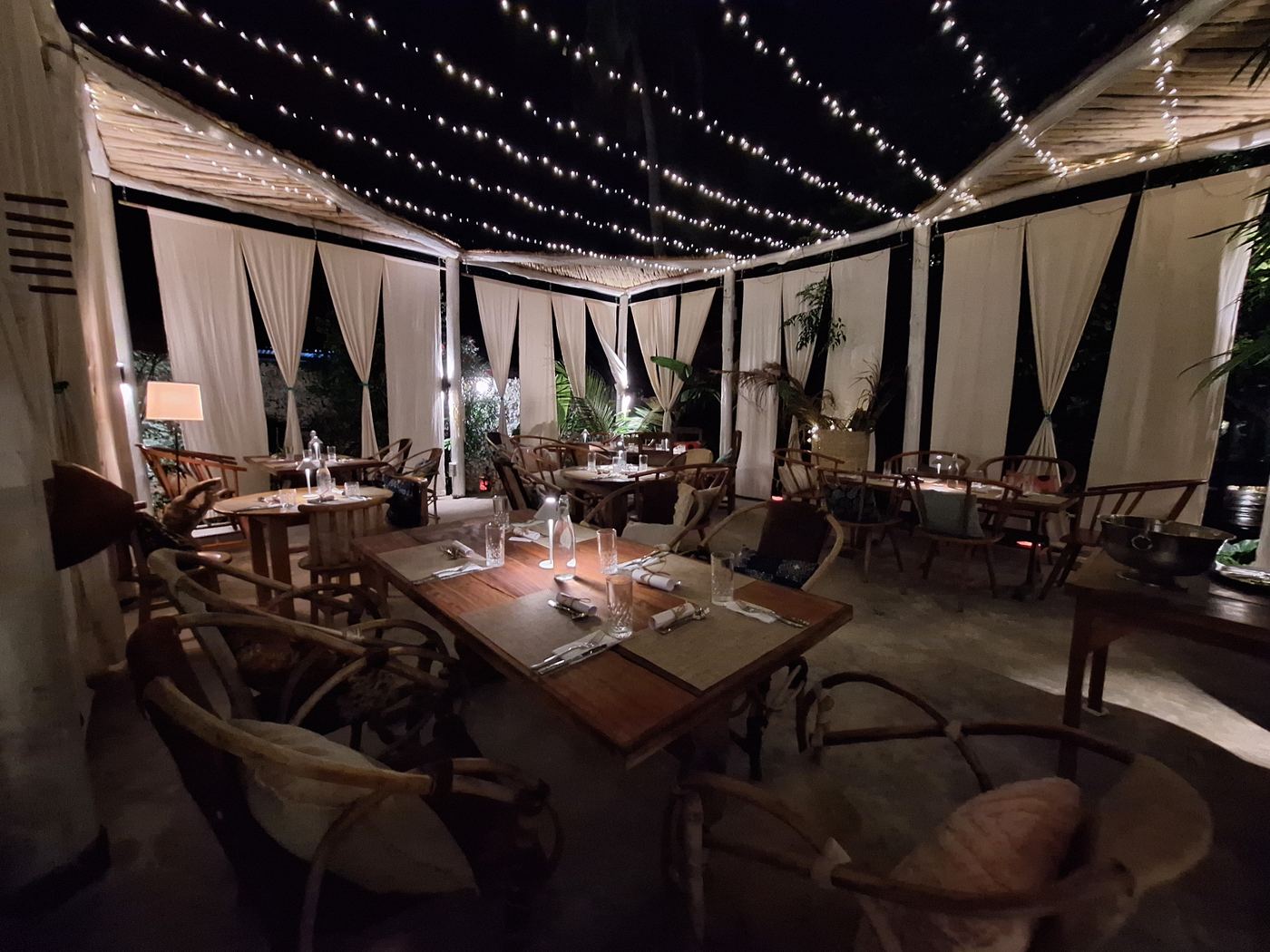 Restaurant tables set for dinner under string lights and white curtains